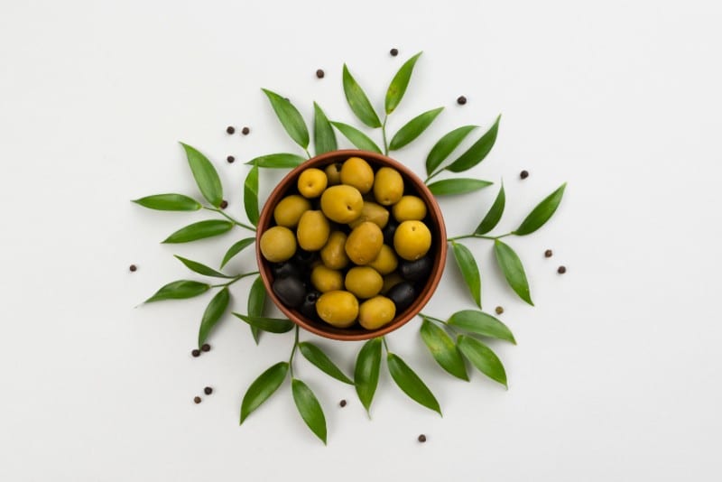 Bowl of green and black olives with surrounding leaves on the table